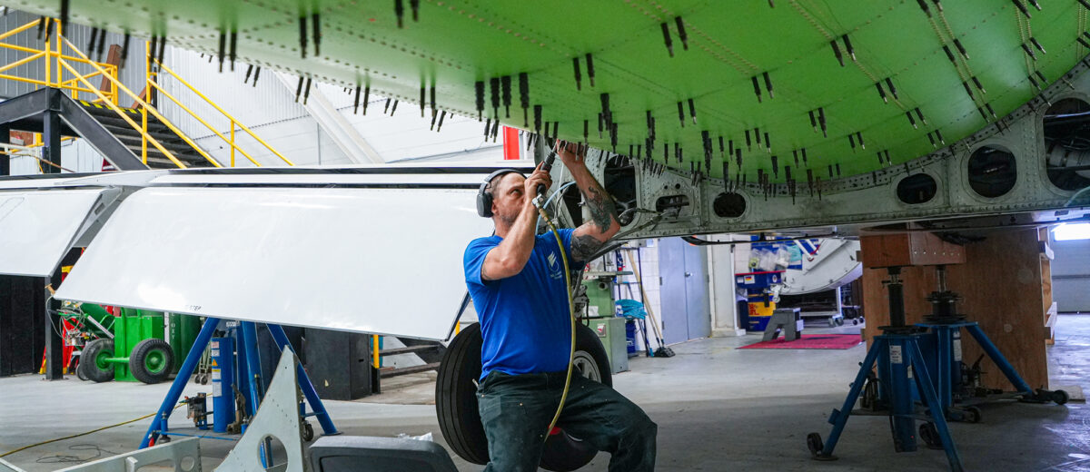 A photo of a mechanic standing underneath the underside of an airplane, fastening bolts on a bright-green section